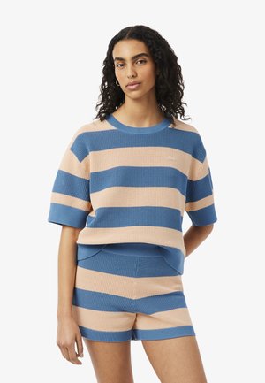 Vrouw draagt een blauw-beige gebreide top met brede strepen en korte mouwen en bijpassende korte broek, staand tegen een effen witte achtergrond.