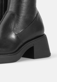 Botte de cheville en cuir noir avec un bout carré, fermeture éclair sur le côté et un talon chunky anglé. Présente une texture lisse et des détails de couture minimaux.