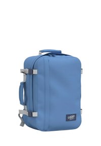 Zaino blu con una forma rettangolare, realizzato in tessuto testurizzato, dotato di una maniglia superiore e spallacci laterali, contrassegnato da un logo.