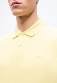 Κοντινό πλάνο ενός ατόμου που φοράει ανοιχτοκίτρινη μπλουζάκι πόλο με γιακά, δύο λευκά κουμπιά και κεντημένο το λογότυπο "BOSS" στο στήθος.