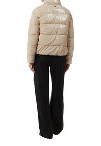Beżowa kurtka puchowa z błyszczącą, pikowaną fakturą, wysokim kołnierzem oraz detalem logo z tyłu, zestawiona z czarnymi spodniami o szerokich nogawkach.