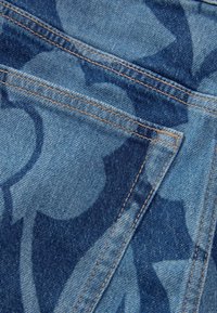 Denim tkanina u svijetloplavoj boji s tamnoplavim cvjetnim uzorkom. Ima vidljive šavove i detalje džepova. Glatka tekstura.