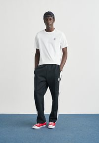 Λευκό μπλουζάκι με κοντά μανίκια και λογότυπο, μαύρο παντελόνι με λευκές ρίγες στα πλάγια και κόκκινα αθλητικά παπούτσια. Το μοντέλο στέκεται σε απλό φόντο.