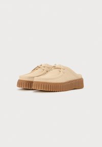Beige suede slip-on shoes with a rounded toe, featuring a ridged rubber sole and thin laces across the front.