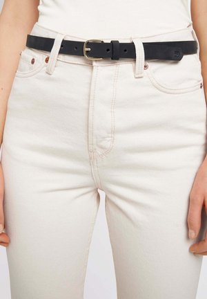 Ceinture en cuir noir avec une boucle en or rectangulaire. Portée par-dessus un jean taille haute beige clair avec des coutures subtiles et une texture lisse.