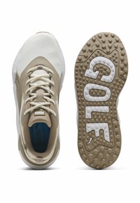 Ζευγάρι μπεζ και λευκά παπούτσια γκολφ Puma, εμφανιζόμενα από πάνω και από τη σόλα, με υφή πρόσφυσης και εμφανές λογότυπο Puma στη σόλα και τη γλώσσα.