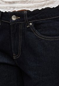 Ζωνη με σκούρο μπλε τζιν, με ορατό κουμπί, μπροστινή τσέπη και λευκή δαντελένια μπλούζα που είναι ελαφρώς βιδωμένη πάνω από τη ζώνη.