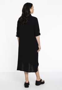 Schwarzes Kleid aus strukturiertem Stoff, mit aufgerollten Ärmeln, seitlichen Schlitzen und einer Länge bis zur Mitte der Wade, kombiniert mit schwarzen Plateauschuhen.