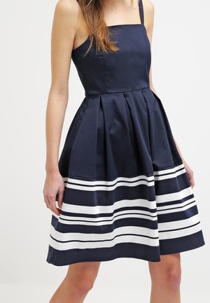 Robe sans manches bleu marine avec décolleté carré et jupe plissée ornée de rayures blanches horizontales près de l'ourlet.