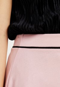 Musta ribattu toppi näkyy osittain vaaleanpunaisen hameen yltä, jossa on musta vyötäröyksityiskohta ja sileä rakenne.