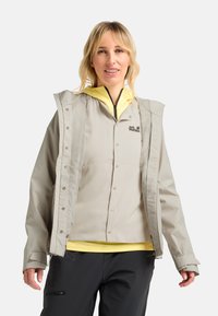 Hellgraue wasserdichte Jacke mit Kapuze, Druckknöpfen und Mesh-Details. Über einem gelben Hemd getragen, kombiniert mit dunklen Hosen.