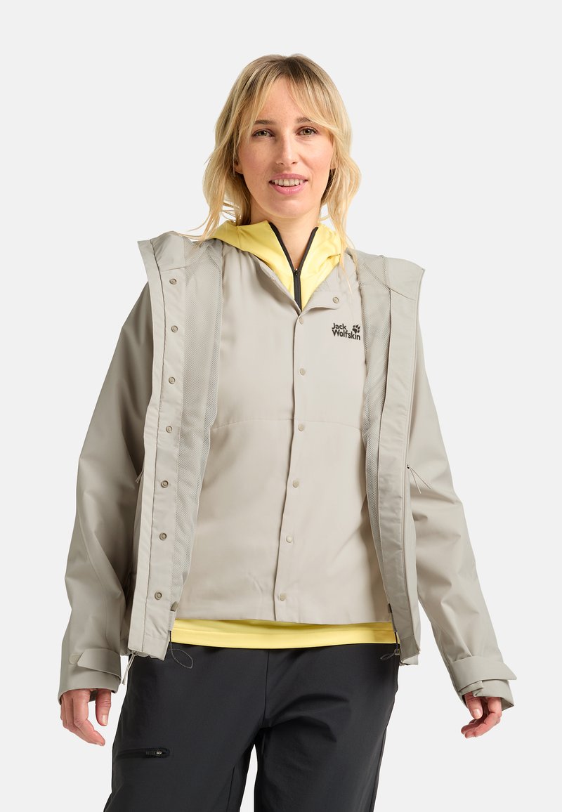 Hellgraue wasserdichte Jacke mit Kapuze, Druckknöpfen und Mesh-Details. Über einem gelben Hemd getragen, kombiniert mit dunklen Hosen.