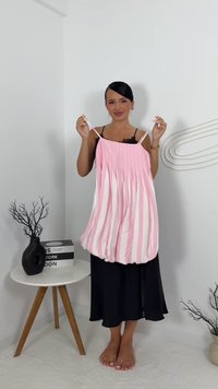 Frau mit dunklen Haaren hält ein rosa-weiß gestreiftes, plissiertes Kleid und steht barfuß auf einem weißen Teppich in einem minimalistischen Raum.