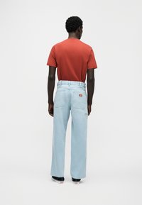 Κόκκινο βαμβακερό T-shirt και ανοιχτό μπλε τζιν με ευθύ κόψιμο, με πίσω τσέπες και μικρό λογότυπο. Μαύρα αθλητικά παπούτσια ορατά.