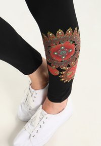 Černé legíny s barevným květinovým vzorem na lýtkách, se červenými, zelenými a modrými akcenty, kombinované s bílými teniskami.