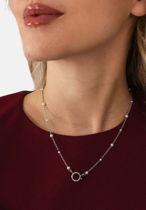 Nahaufnahme einer Person, die eine silberne Kettenhalskette mit kleinen Perlen und einem runden Verschluss trägt, kombiniert mit einem dunkelroten Oberteil.