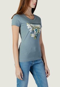 Svijetloplava majica kratkih rukava s trokutastim cvjetnim motivom i ukrasnim znakom upitnika u sredini.