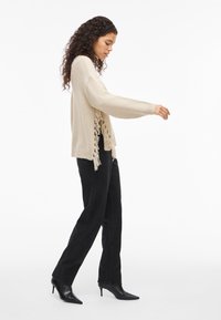 Γυναίκα που περπατάει φορώντας ένα μπεζ πουλόβερ με φράντζες, μαύρα τζιν ίσιων ποδιών και μαύρες γόβες με尖尖 έκτροφό προς τα πάνω σε λευκό φόντο.