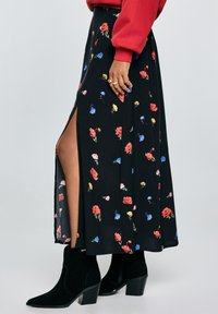 En person har en rød sweater på og et blomsterprintet sort nederdel med en slids op til låret, parret med sorte ankelstøvler, stående mod en lys baggrund.