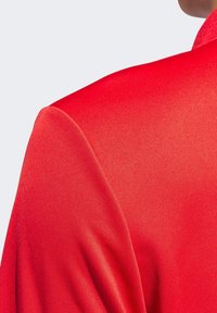 Rød jakke i et glat, skinnende stof med struktur i skuldrene og revers, der fremhæver dens skræddersyede design og livlige farve.