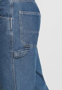 Farkut sinisellä sävyllä ja hieman haalistuneella tekstuurilla, joissa on suuri takatasku ja kontrastiset ompelut.