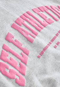 Grå sweatshirt med fet, upphöjd rosa text som läser "SJÄLVAWARENESS" med en något texturerad yta. En anmärkningsvärd detalj inkluderar en extra text nedanför.