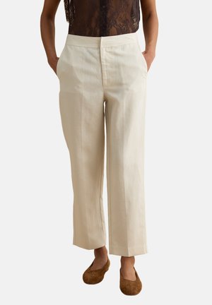 Mujer con pantalones crema de talle alto y pierna recta, con bolsillos laterales y zapatos planos de ante marrón, de pie frente a un fondo liso.