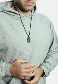 Schwarze Hundemarke-Halskette mit kompliziertem Muster, getragen über einem grauen Hoodie. Sie verfügt über eine lange schwarze Kette und einen strukturierten, rechteckigen Anhänger.