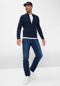 Mørkeblå zip-up jakke i blødt stof, parret med en hvid crew neck skjorte, mørke jeans og hvide sneakers. Inkluderer en mørkeblå cap med lyseblå accent.