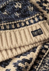 Gebreide muts in marineblauw en crème, met geometrische patronen, geribbelde rand en een gestructureerd oppervlak. Voorzien van een blauw label met witte tekst.