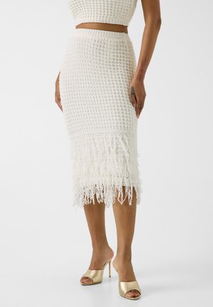 Femme portant une jupe midi blanche en crochet avec des franges au bas et des sandales dorées à talons hauts ouvertes sur les orteils, debout sur un fond uni.