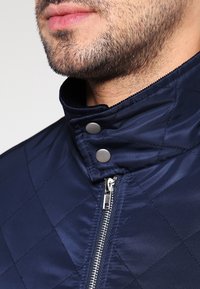 Lähellä otettu kuva miehen alhaalta kasvoista, joissa on hoidettua partaa, ja hänellä on yllään tummansininen kulutettu takki, jossa on vetoketju ja napillinen kaulus.