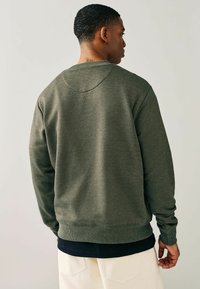 Olivgrön sweatshirt med rund halsringning, ribbade ärmslut och nederdragskant. Mjuk textur, avslappnad passform, bärs över en mörk skjorta, visad från baksidan.