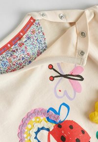 Tricou din bumbac crema cu imprimeuri colorate de fluturi și buburuze, volane decorative și guler interior cu model floral. Butoniere cu capsenă incluse.