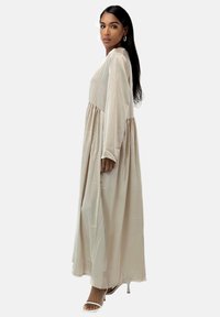 Lang beige kjole med lange ærmer og en taljesamling. Glat, letvægtigt stof falder til ankellængde. Kombineret med hvide hælede sandaler.