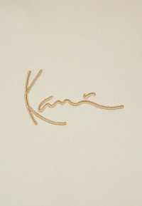 Beige stoff med en myk tekstur, prydet med en brodert signatur i gyllen tråd, som viser buede linjer og et flytende design.