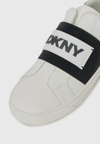 Sneaker bianco realizzato in materiale liscio, con una banda elastica nera decorata con il logo "DKNY". Suola in gomma con dettagli texturizzati.