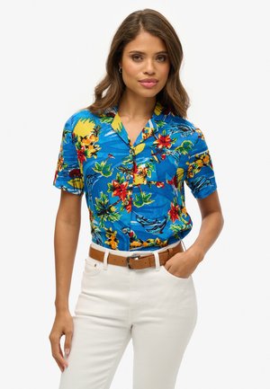 Une chemise bleue à manches courtes ornée de fleurs jaunes et rouges éclatantes, de feuilles tropicales, et dotée d'un bouton devant. Ceinture avec un pantalon de couleur claire.