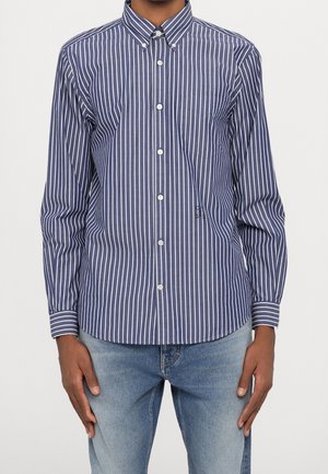 Man draagt een lange mouwen donkerblauw overhemd met witte verticale strepen en lichtblauwe spijkerbroek, staat met ontspannen handen langs het lichaam.