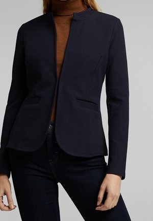 Kvinde iført en tætsiddende sort blazer over en brun rullekrave og mørke jeans, stående mod en neutral baggrund.