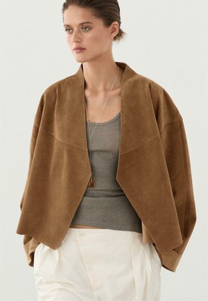 Młoda kobieta ubrana w luźną brązową zamszową kurtkę na szarym prążkowanym topie na ramiączkach i białych spodniach z wysokim stanem, stoi na prostym tle.