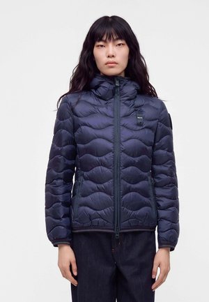 Woman with long dark hair wearing a navy blue quilted jacket with high collar and front zipper, standing against a plain white background.
