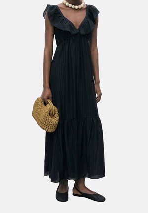 Kvinde iført en sort maxikjole uden ærmer, stor perlebesat halskæde, bærende en vævet tanfarvet håndtaske og sorte flade sko.