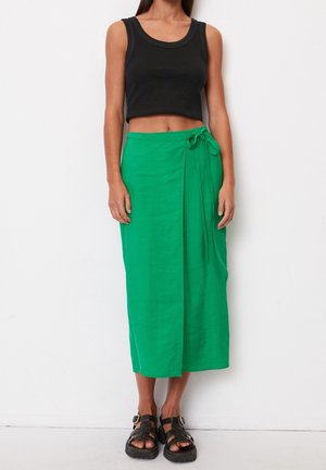 Mujer vistiendo una camiseta negra corta, una falda midi verde con nudo lateral y sandalias negras gruesas, de pie contra un fondo blanco.