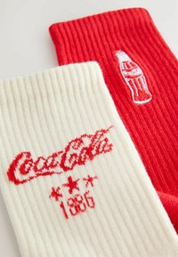 Valged ja punased triibulised sokid, millel on broderieeritud Coca-Cola logo ja pudel. Tekst sisaldab "Coca-Cola" ja "1886" punases.