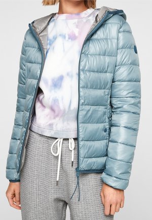 Žena nosící světle modrou prošívanou péřovou bundu s kapucí, mikinu s batikou v bílé, fialové a modré barvě a šedé tepláky s waffle pletením a stahovací šňůrkou.