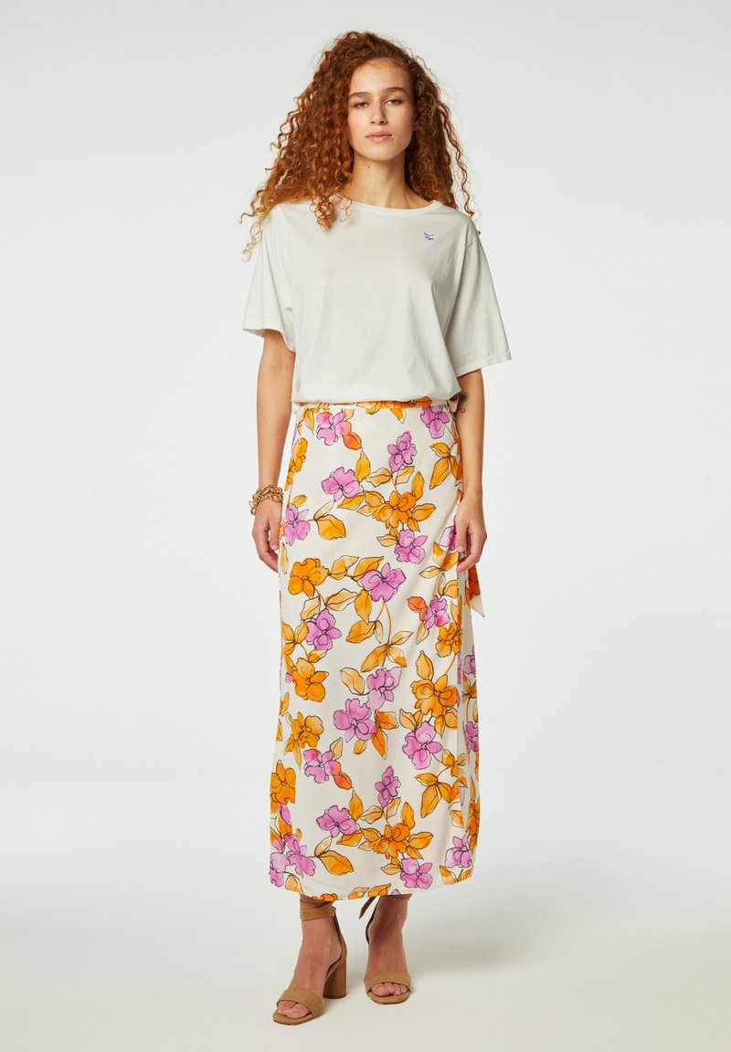 Lichtgrijze shirt met korte mouwen, gecombineerd met een luchtige witte rok met levendige oranje en roze bloemenpatronen. Beige blokhak sandalen.