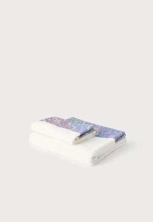 Dos toallas blancas dobladas con bordes estampados en azul y morado de motivo cachemir sobre un fondo liso gris claro.