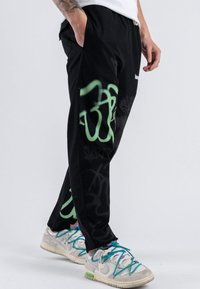 Pantaloni felpati neri con design in stile graffiti verde. Presentano una vita elastica e tasche laterali. Indossati con sneakers grigie e lacci colorati.