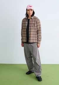 Chemise à carreaux marron boutonnée, t-shirt noir, pantalon en velours côtelé gris, casquette rose avec texte rouge, baskets noires, debout sur un sol vert.